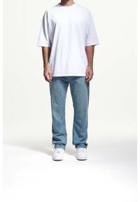 Bílé přepravné tričko s krátkým rukávem, kombinované se světle modrými džíny a bílými teniskami, stojící na jednolitém pozadí.