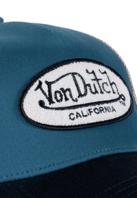 Gorra verde azulado con visera azul marino oscura, que presenta un parche ovalado bordado en negro con el texto "Von Dutch CALIFORNIA" en letras blancas.