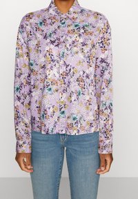Camicia a maniche lunghe floreale di colore viola chiaro con motivi di fiori multicolori. Presenta un colletto e una chiusura con bottoni sul davanti, abbinata a jeans blu.