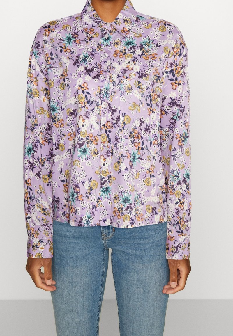 Langärmliges, blumiges Hemd in Helllila mit mehrfarbigen Blumenmustern. Verfügt über einen Kragen und eine Knopfleiste, kombiniert mit blauen Jeans.