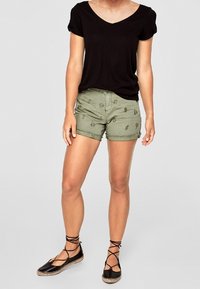 Haut noir à manches courtes en V associé à un short vert olive avec un motif ananas subtil, porté avec des espadrilles noires à lacets.