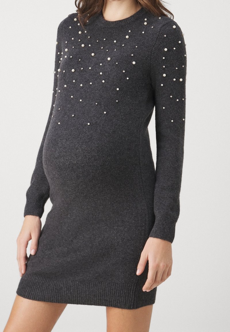 Femme enceinte portant une robe en maille gris foncé décorée de petites perles blanches sur la poitrine et les manches, debout, mains relâchées le long du corps.