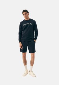Granatowy sweatshirt z napisem "COSTA DEL SOL", w zestawie z dopasowanymi szortami, kremowymi sneakersami i białymi skarpetkami. Prosty, casualowy design i krój.