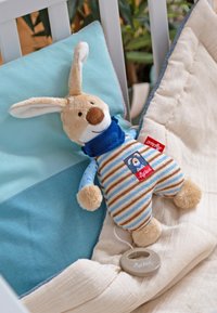Jouet lapin en peluche vêtu d'une salopette rayée et d'une écharpe bleue, posé sur une couverture beige et un coussin bleu dans un berceau blanc avec des plantes vertes en arrière-plan.