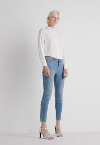 Fehér gombos ing, világoskék skinny farmernadrág és ezüst magas sarkú szandálok. Simább anyag, testhez simuló dizájn és diszkrét részletek a ruházaton.