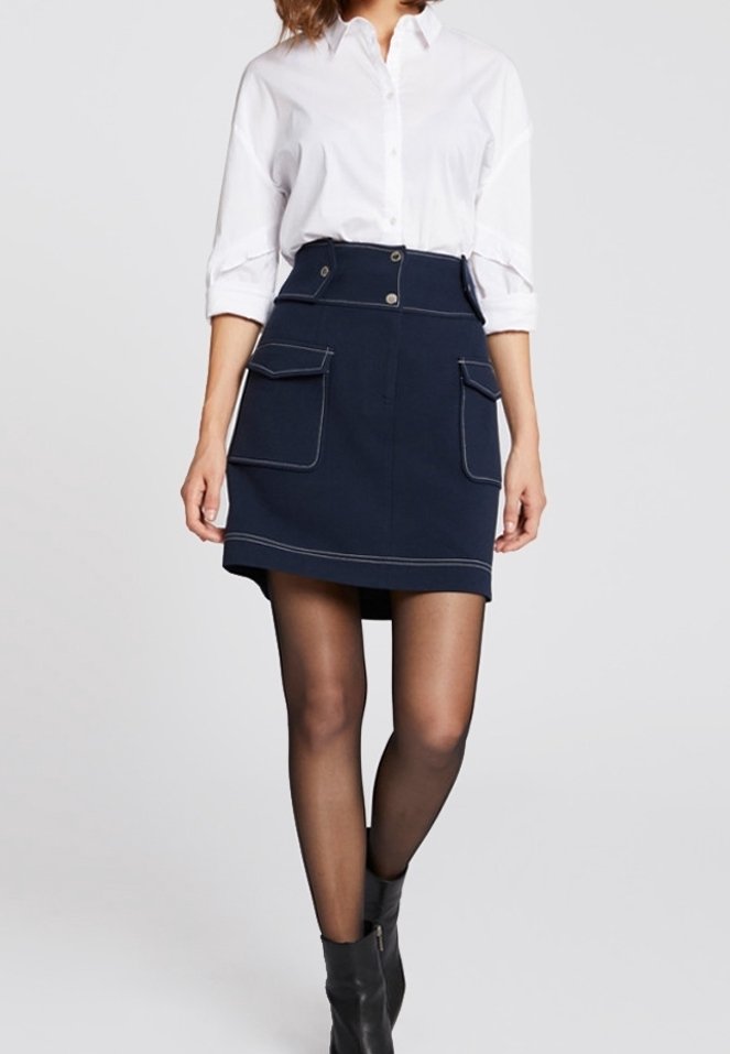 Falda azul marino con dos bolsillos frontales de parche, costuras en contraste y una cintura alta. Combinada con una camisa blanca de botones y botas de tobillo negras.