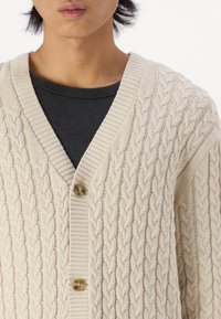 Lys beige kabelstrikket cardigan laget av strikket stoff, med V-hals, ribbestrikkede kanter og to treknotter foran.