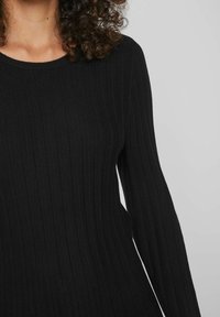 Persona dai capelli ricci che indossa un maglione a maniche lunghe a coste nere su uno sfondo grigio chiaro uniforme.