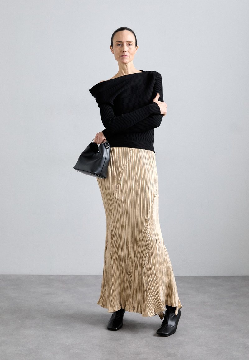 Pull noir à côtes, jupe plissée dorée métallique, sac à main structuré noir et bottines pointues noires sur fond gris.