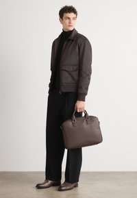 Veste bomber marron foncé avec deux poches avant, portée sur un col roulé noir. Le modèle tient une mallette en cuir assortie. Pantalon et chaussures noirs.