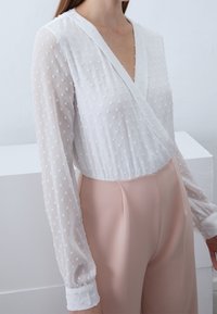 Blusa blanca con lunares texturizados, escote en V y mangas largas transparentes, combinada con pantalones rosa de tiro alto. Acabado de tela suave.