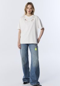 T-shirt bianco a maniche corte con catene decorative e ciondoli; abbinato a jeans a gamba larga blu chiaro con un'etichetta verde su una catena.