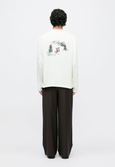 Camisa branca de manga comprida com um gráfico de um esquiador e montanhas nas costas, combinada com calças de perna larga castanho escuro. Texto próximo ao design diz "WONDERLAND."