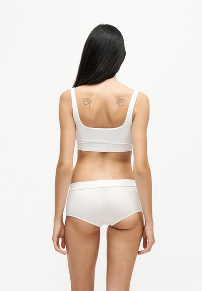 Femme aux longs cheveux noirs portant un soutien-gorge et une culotte blancs, montrant deux tatouages de l'Œil d'Horus sur le haut du dos sur un fond blanc uni.