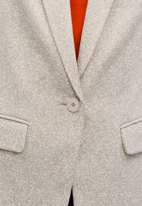 Lichtgrijze blazer van gestructureerde stof, met een enkele knoopsluiting en twee voorzakken. Daaronder is een oranje overhemd zichtbaar.