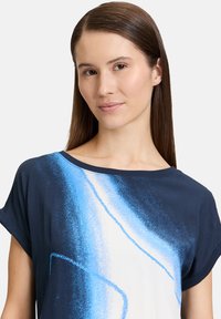 Granatowa bluzka z krótkim rękawem z abstrakcyjnym wzorem w niebiesko-białe motywy. Miękka tkanina o luźnym kroju i szerokim dekolcie.