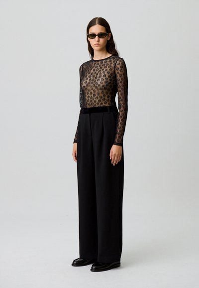 Femme portant un pantalon large noir, un haut en dentelle florale transparente à manches longues, des mocassins noirs et des lunettes de soleil sombres, se tenant devant un fond uni.