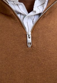 Maglione marrone con cerniera, superficie testurizzata, con una cerniera argentata e una camicia bianca a collo con sottili righe sotto.