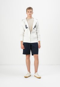 Hvit zip-up hettegenser med marineblå detaljer, beige t-skjorte, marineblå shorts og lyse sneakers. Glatt stoff, atletisk passform, minimalistisk design.