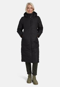 Melns garš pūkains mētelis ar kapuci, ar rāvējslēdzēju, sānu kabatām un horizontālu quiltēšanu. Uzvilkts virs tumši zaļām biksēm un zābakiem.