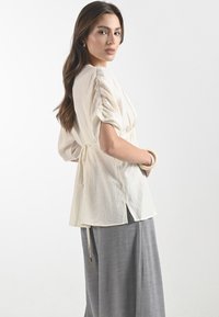 Il modello indossa un top color crema chiaro con maniche arricciate, abbinato a una gonna grigia, mostrando un look semplice ed elegante su uno sfondo neutro.