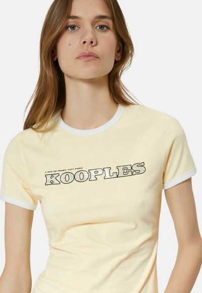 Young woman wearing pale yellow t-shirt with white trim and black "KOOPLES" text across the chest on a plain white background.