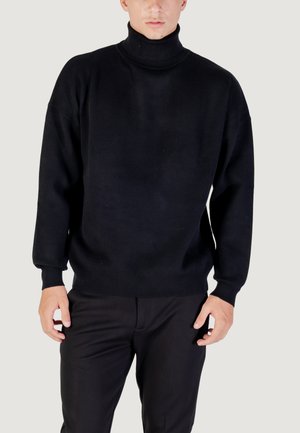 Maglione nero a collo alto in morbido tessuto a maglia con spalle scese e polsi a coste, abbinato a pantaloni scuri, esposto su uno sfondo chiaro.