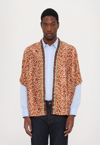 Chaqueta estilo kimono de textura marrón y crema con un patrón; presenta un ribete en contraste en verde y marrón oscuro, llevada sobre una camisa azul clara.