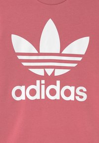 T-shirt en coton rose arborant un grand logo trefoil blanc d'Adidas et des rayures horizontales en dessous. Texture douce avec un col rond.