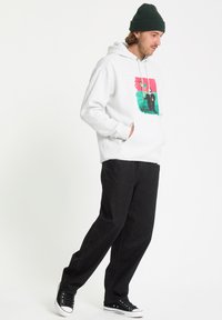 Un hoodie gris avec un imprimé graphique coloré à l'avant, associé à un pantalon noir à jambes droites et des baskets noires. Bonnet en laine sur la tête.