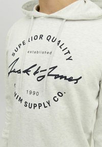 Lys grå hættetrøje med snoretræk, der har et stort sort trykt logo, som står "Superior Quality" og "Jack & Jones Denim Supply Co."