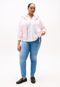 Roze zip-up hoodie over een witte button-up shirt, gecombineerd met lichtblauwe skinny jeans en zwarte slip-on schoenen. Gladde, casual stoffen.