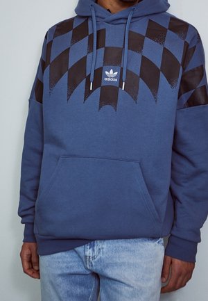 Muž nosící modrou mikinu Adidas s černým geometrickým vzorem a klokaní kapsou, v kombinaci se světle modrými džínami.