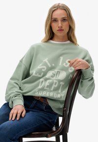 Sudadera de cuello redondo verde claro con texto gráfico en blanco, combinada con jeans azul oscuro, se encuentra sobre una silla de madera. El texto incluye "EST. 1985" y "SUPERDRY."