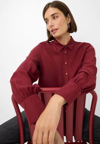 Burgundy button-up shirt with a classic collar and gathered sleeves, paired with a modern red chair. Smooth fabric texture, tailored design.