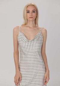 Robe en satin blanche à pois noirs, décolleté plongeant, fines bretelles et silhouette ajustée. Texture lisse et accents de design minimalistes.