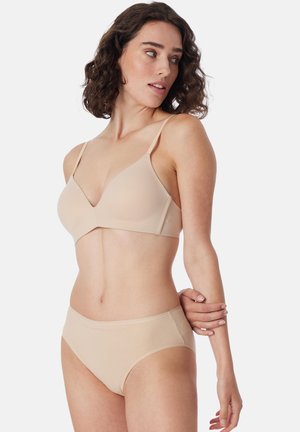 Vrouw met krullend donker haar, een beige draadloze beha en bijpassend ondergoed, kijkt naar links tegen een effen achtergrond.