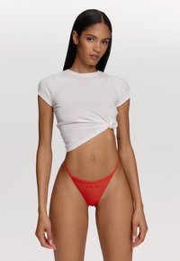 Tanga rouge avec le texte « KISS ME », fines bretelles et couverture minimale, associé à un haut blanc côtelé à manches courtes avec un design noué.