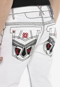 Weiße Jeans verfügen über schwarz-rote gestickte Designs auf den Gesäßtaschen, mit kontrastierendem Nähen und markanten Taschenformen.