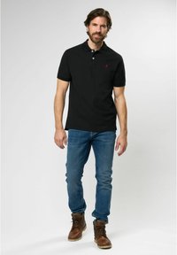 Black polo shirt with a collar and short sleeves, accented by a small red logo. Worn with blue jeans and brown lace-up boots.