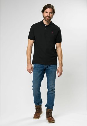 Sort polo skjorte med krave og korte ærmer, accentueret af et lille rødt logo. Båret med blå jeans og brune snøresko.