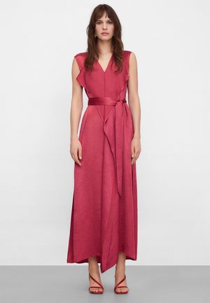 Kobieta ubrana w bezrękawową, głęboko różową sukienkę kopertową z paskiem i czerwone sandały na obcasie, stojąca na tle jednolitej białej ściany.