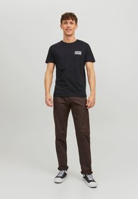 Schwarzes Baumwoll-T-Shirt mit weißem "JACK & JONES"-Logo auf der Brust, kombiniert mit braunen Hosen und schwarzen High-Top-Sneakers.