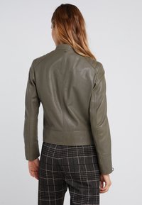 Oliivinvihreä nahkatakki, jossa on kihartava muotoilu, seisova kaulus ja ompeluyksityiskohtia, yhdistettynä ruutukuosisiiin housuihin.