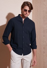 Camisa azul marino con cuello mao, mangas largas arremangadas en los puños, hecha de tela texturizada. Combinada con pantalones de color claro.