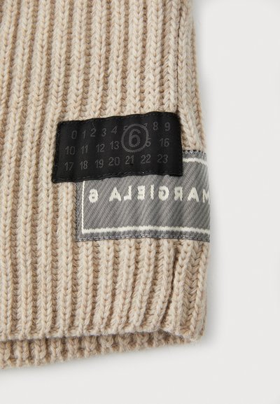 Chapeau en tricot beige avec une texture côtelée et une étiquette de soin noire comportant des chiffres et une pièce en tissu avec logo cousue le long de la couture.