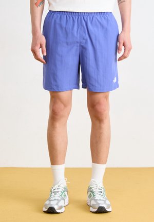 Person trägt blaue Sportshorts, weiße Socken und grau-weiß-grüne Laufschuhe und steht auf einem gelben Boden mit hellem Hintergrund.