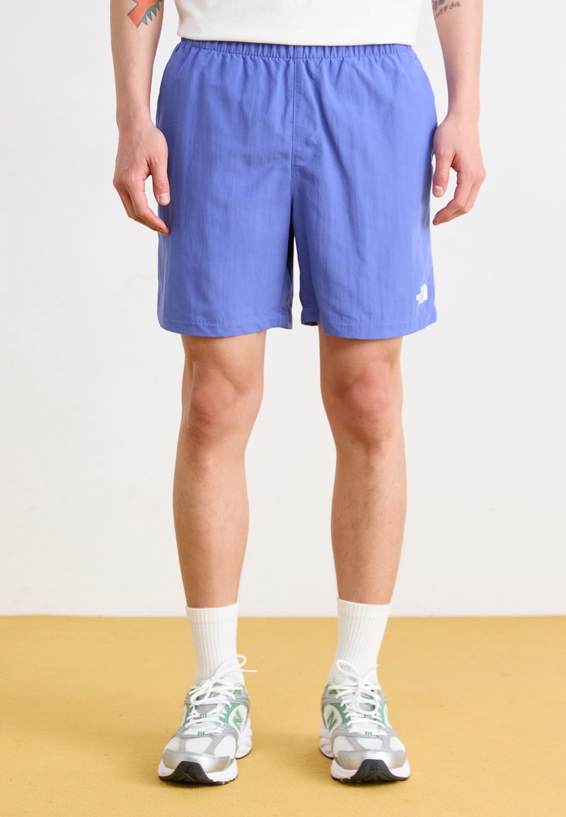Personne portant un short de sport bleu, des chaussettes blanches et des chaussures de course gris-blanc-vert, debout sur un sol jaune avec un fond clair.