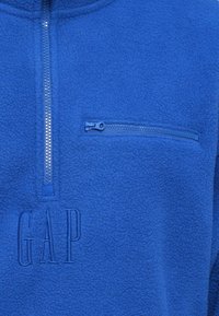 Kék polár pulóver negyedcsíkos cipzárral, hímzett "GAP" logóval és cipzáras mellzsebbel. Puha textúra.