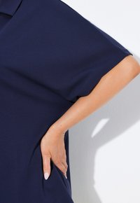 Donkerblauw jurkje met losvallende korte mouwen, gemaakt van een gestructureerde stof. Hand op de heup geplaatst, wat de silhouet van het jurkje accentueert.